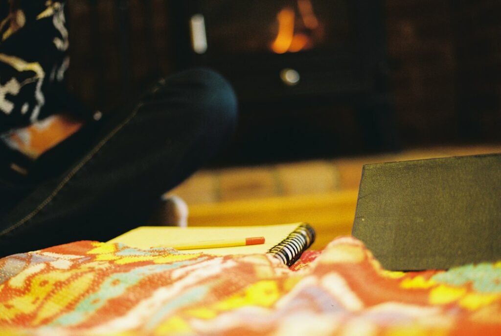 An open notebook and pencil on a patterned throw with a glowing fireplace