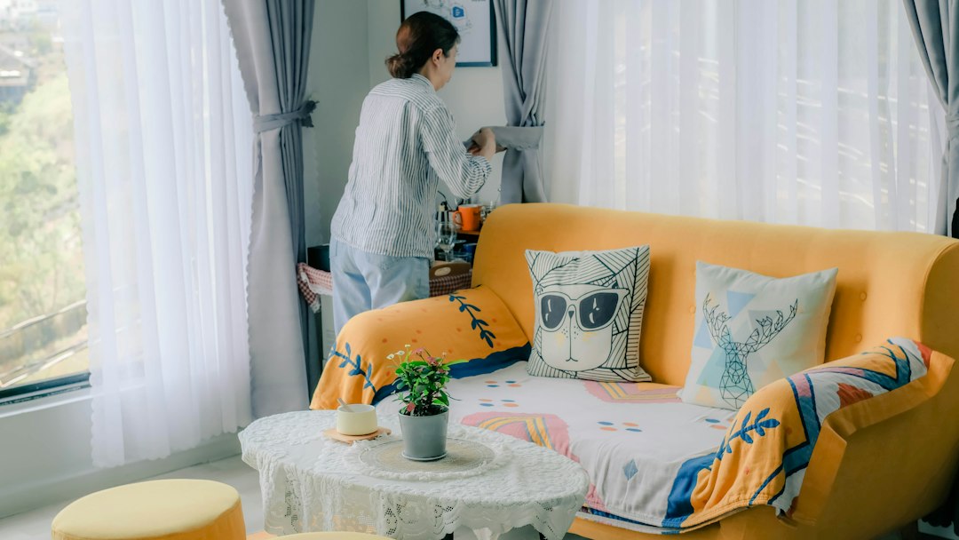 Do Air Purifiers Help With Dust Mites – A woman adjusting curtains in a sunlit living room with a yellow sofa